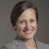 Portrait of a smiling woman in a gray blazer with pearls, likely a professional in the medical field.