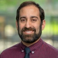 Portrait of a smiling man with a beard, wearing a burgundy checkered shirt and tie.