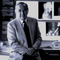 Portrait of a man in a suit, sitting in an office with medical images in the background.
