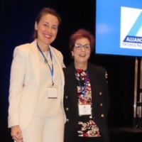 Two women smiling together at a conference, with a blue logo displayed in the background.