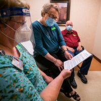 A healthcare worker discusses information with two patients in a clinical setting, all wearing masks and face shields.