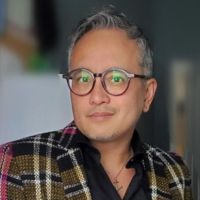 Portrait of a person with short hair and glasses, wearing a plaid blazer.